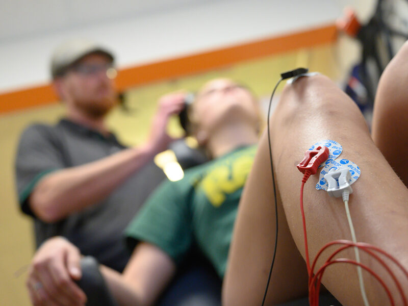 lab member performing electromyography on a participant who sits in a chair with electrodes on their legs. electromyography allows us to record electrical activity directly from active muscles. Through this technology we can determine when muscles are active and how much they are contracting.