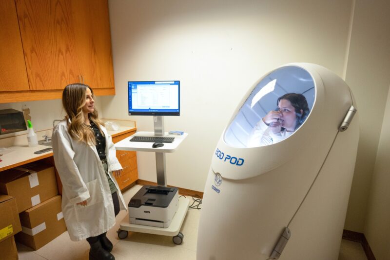 A participant takes a bod pod test while a lab technician examines.