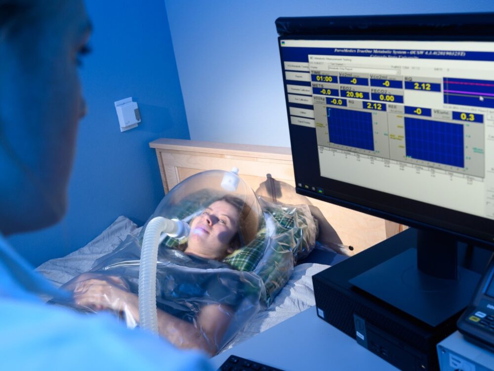 A Resting Metabolic Rate test is conducted as a lab operator examines the test participant, and the participant lays on a table while wearing the resting metabolic rate equipment on their head.