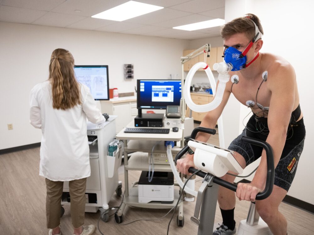 A research participant engages in a training zones determination test while riding a stationary bike connected to tubes and a computer.