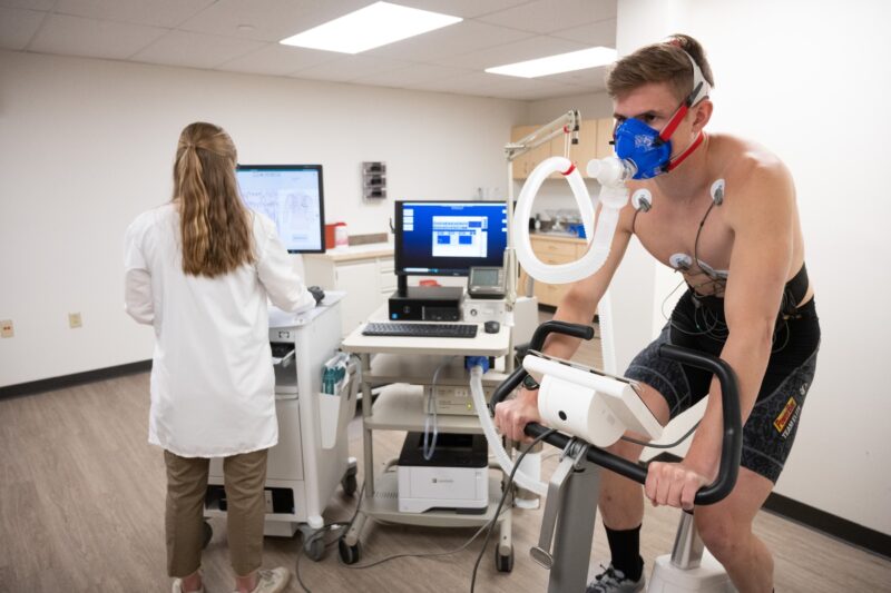 A research participant engages in a training zones determination test while riding a stationary bike connected to tubes and a computer.