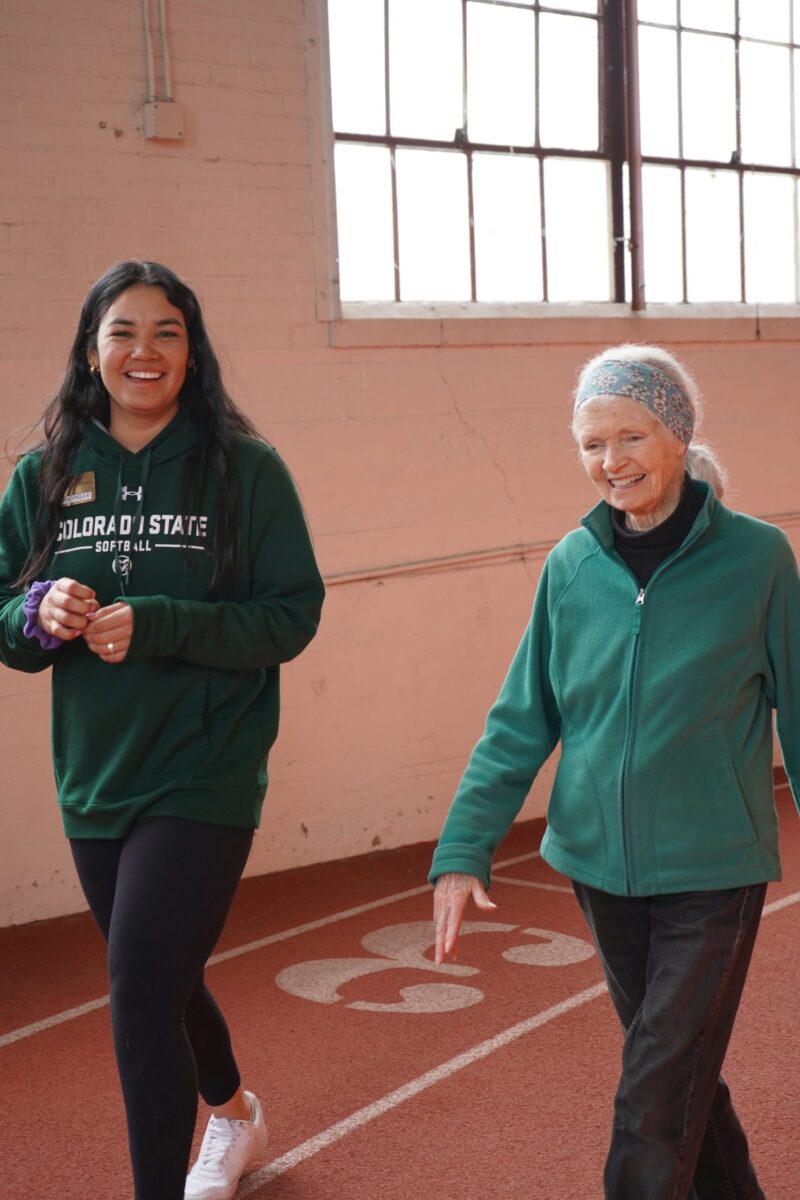 An adult fitness student in a Colorado State University sweatshirt smiles as whe walks with an elderly participant that is also smiling on the track