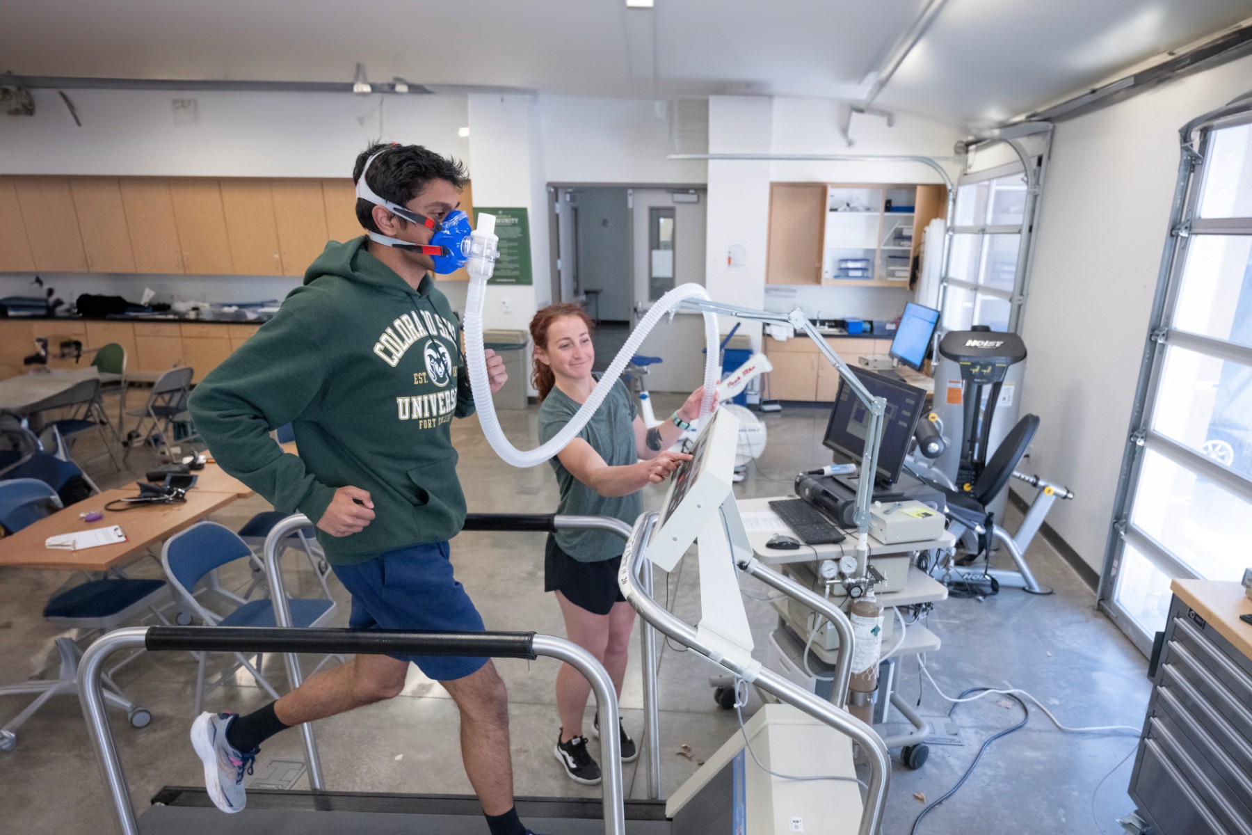 A student runs on a treadmill connected to oxygen tubes while another student reads a machine to determine their oxygen levels.