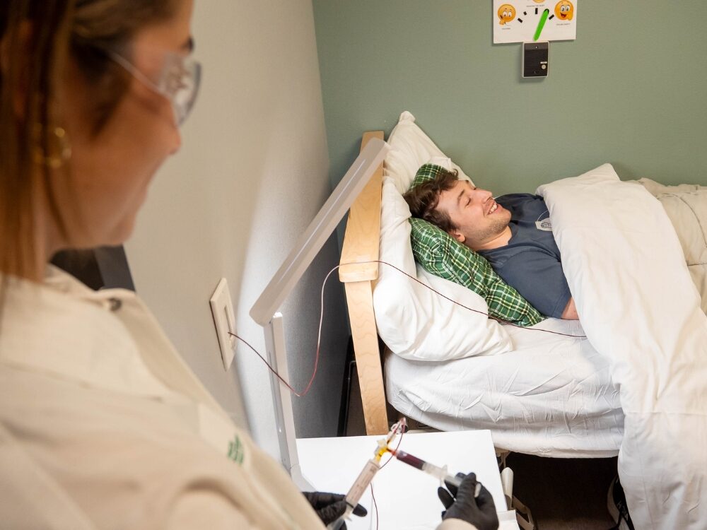 A sleep and metabolism lab participant smiles in a bed while a researcher safely draws blood.