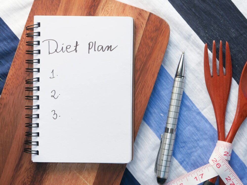 Note book with words `diet plan` on table cloth with pen, wooden spoon and fork nearby