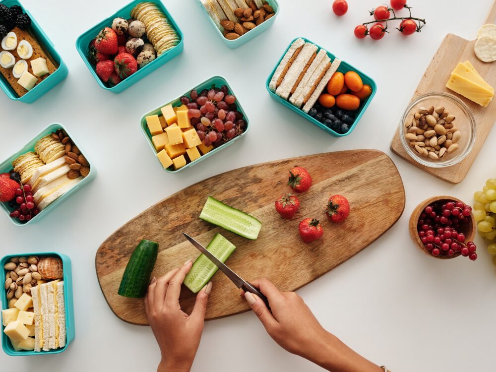 Hands chopping vegetables on wooden chopping board, surrounded by small lunch containers with snacks