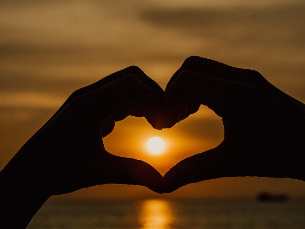 two hands forming a heart shape silhouetted in front of a sunset