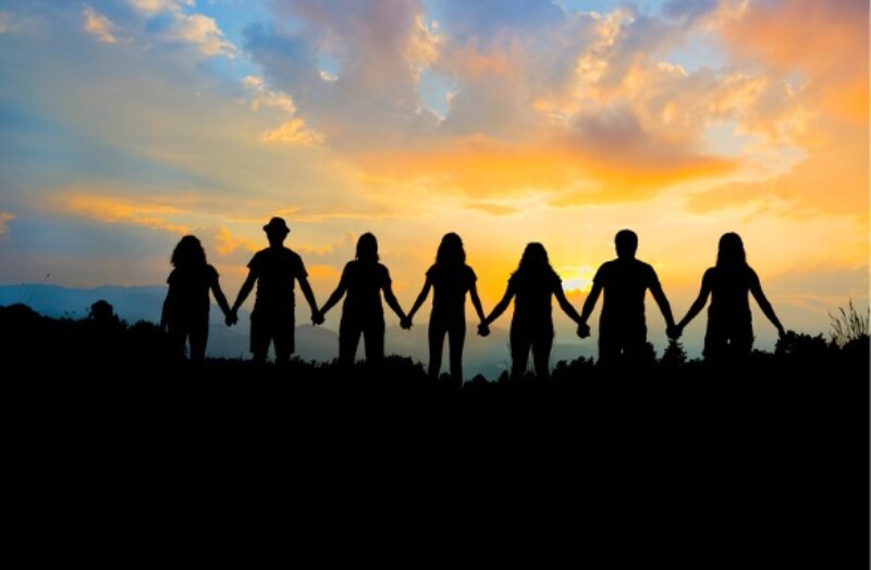 7 people standing in a line, holding hands, silhouetted in front of a sunrise