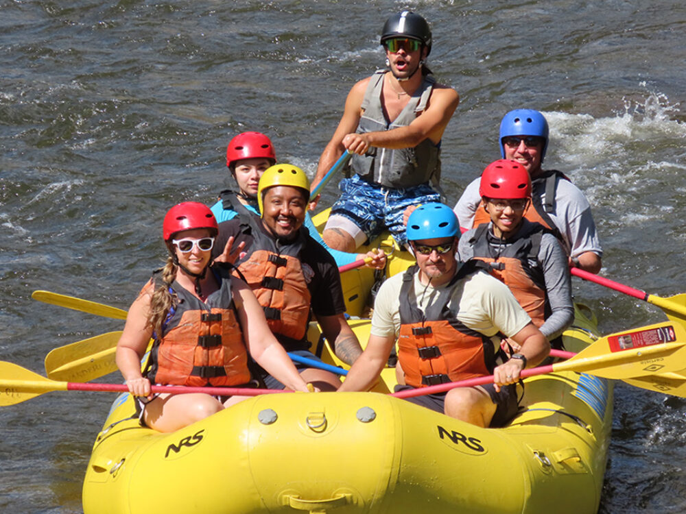 Seven people wearing helmets and life jackets while riding down a river in a bright yellow raft