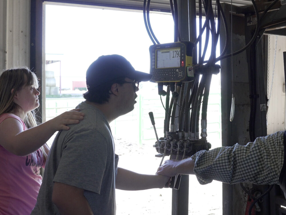 One Ram Scholar pushes machinery levers with instructor guidance, while another Ram Scholar places a hand on their shoulder