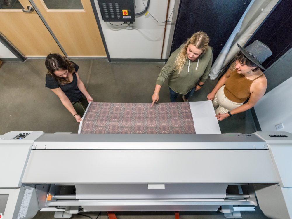 Three students looking at fabric being printed from the fabric printer