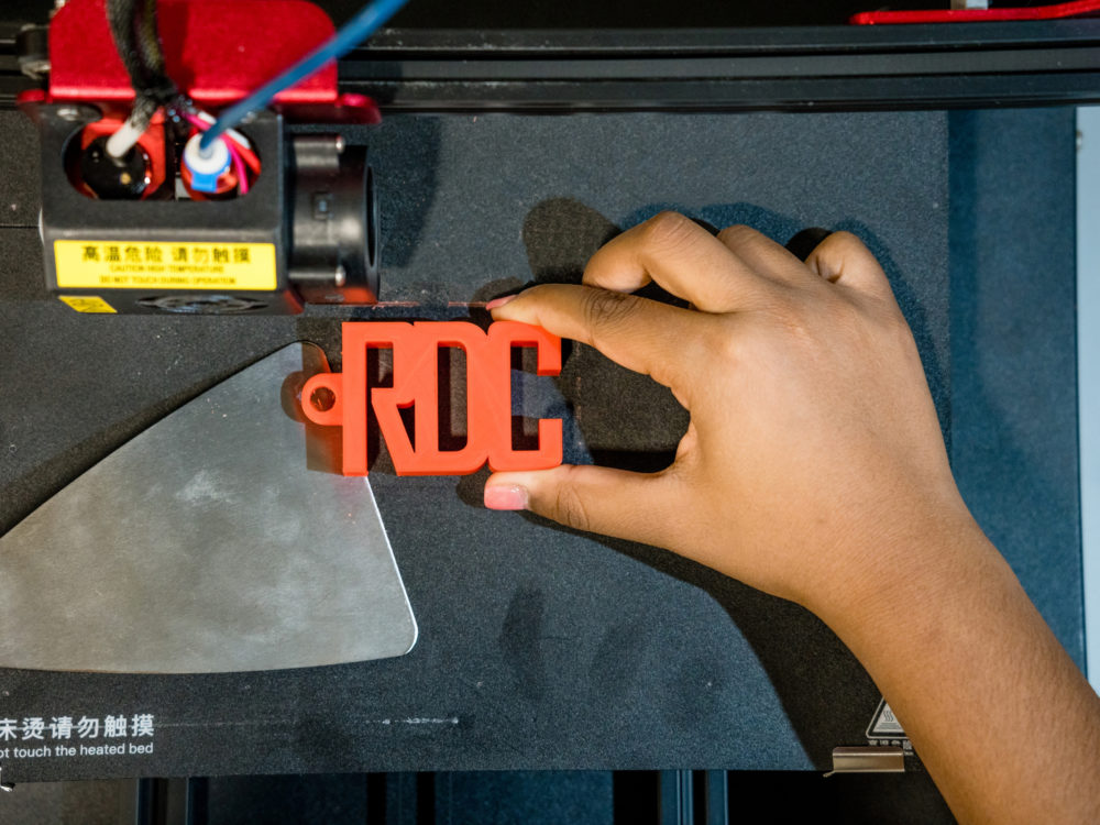 A hand holding an orange 3D printed block of letters reading `RDC`