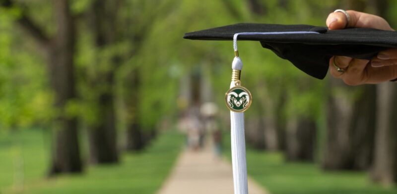 Hand holding graduation cap with rams logo charm and white tassle along Oval path