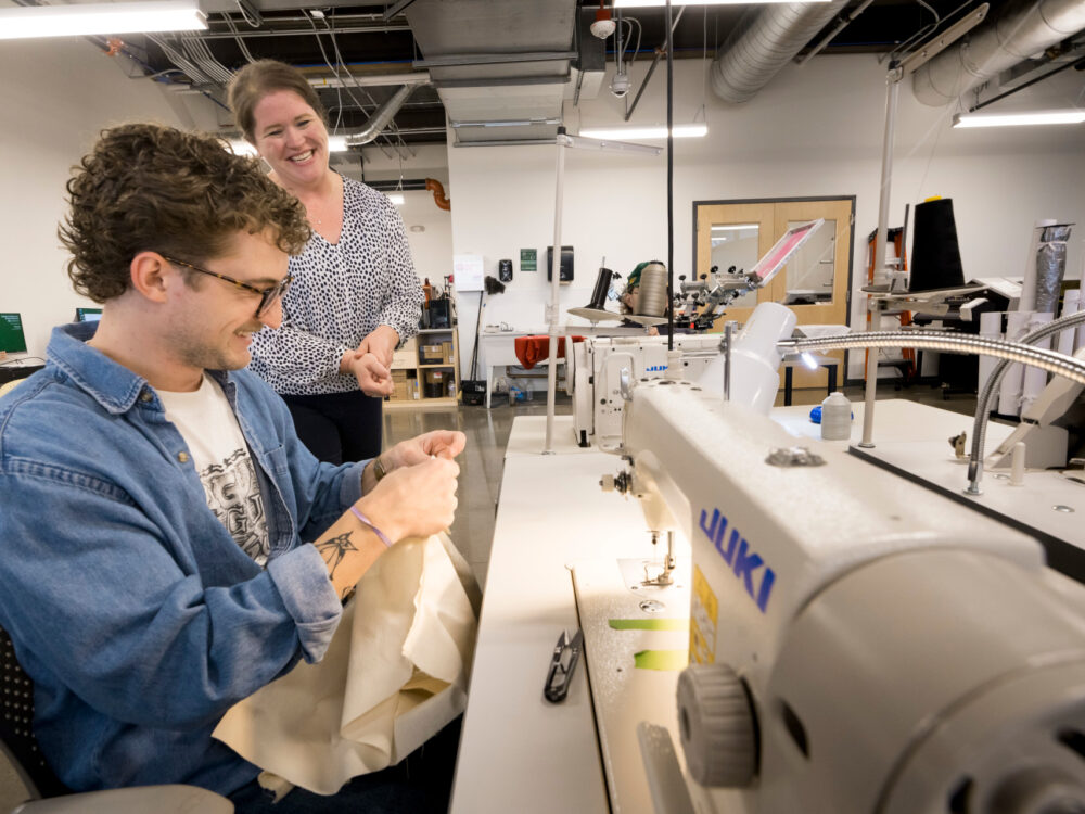 Professor chatting with student who is holding a garment sitting at a sewing machine