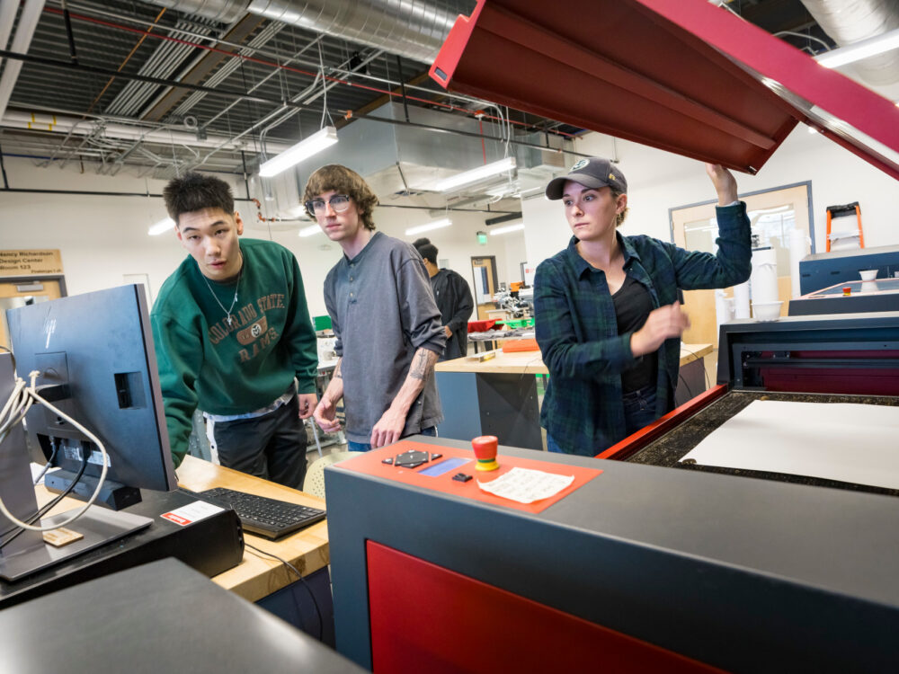 Three students work together on a laser cutter in the Prototype Lab