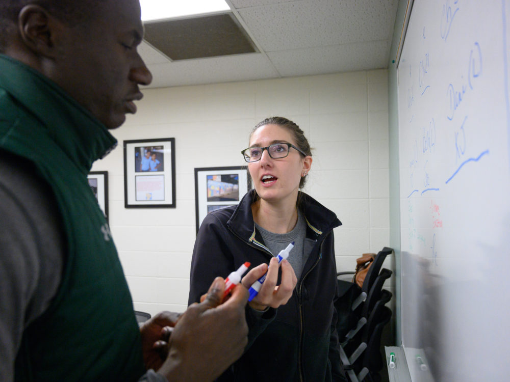 Two students, one presenting as a Black male and one presenting as a white female, discuss as they write on a whiteboard during class