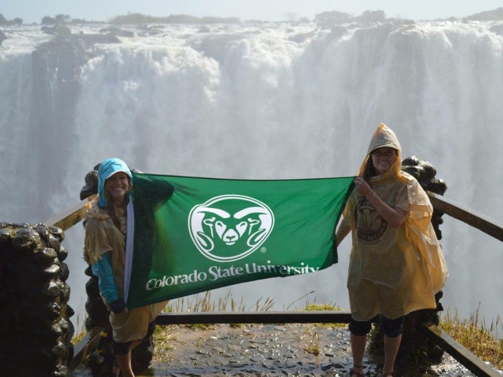 CSU studnets hold up a CSU flag in front of a waterfall in Zambia