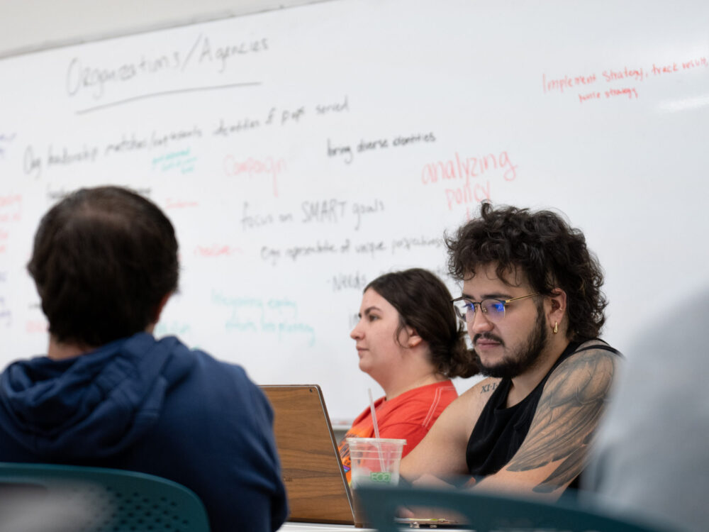 A student reads on their laptop and a whiteboard with notes on it in the background.