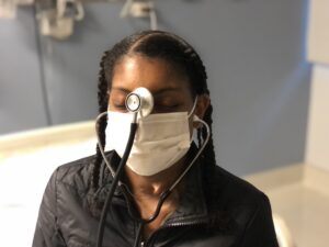 A young woman sits in a hospital setting with a mask and stethoscope. The center of the stethoscope rests between her eyes.