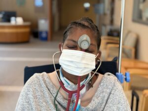 A young woman sits in a wheelchair in a hospital setting with a mask and stethoscope. The center of the stethoscope rests between her eyes.