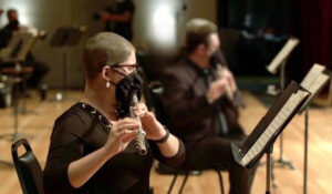 A cancer survivor plays a flute on an indoor stage during a concert or rehearsal, with music stands in front of them.