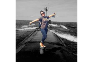 A person balances atop a small structure on a submarine deck, with ocean and conning tower in the background.