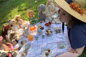 A person sits at a picnic blanket with food and dolls