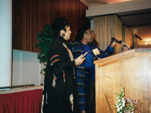 Cheryl speaks at a podium at an event. Next to her is a colleague in an African dress.