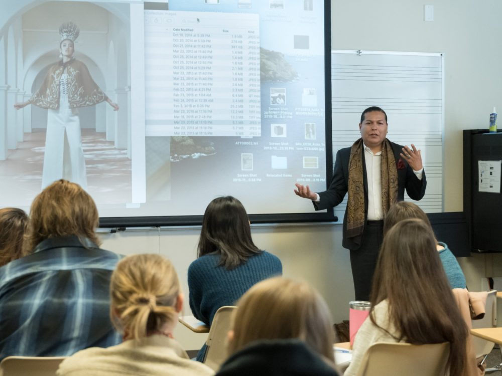 A Native American man stands in front of a class talking with a projected image of a computer screen behind him