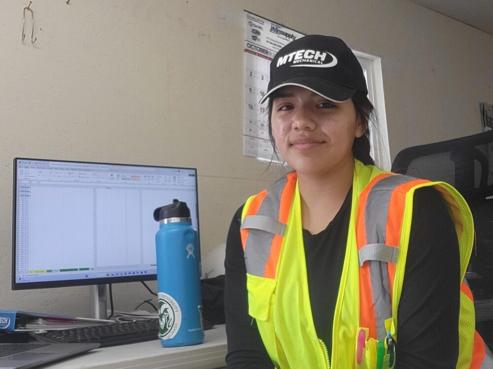 Latina woman in a bright construction vest and MTech cap sits at a computer