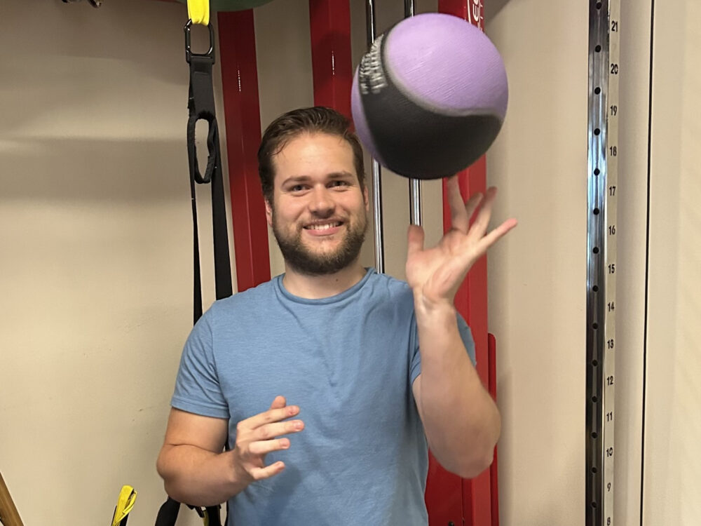 A man in a workout room tosses a blue and purple ball