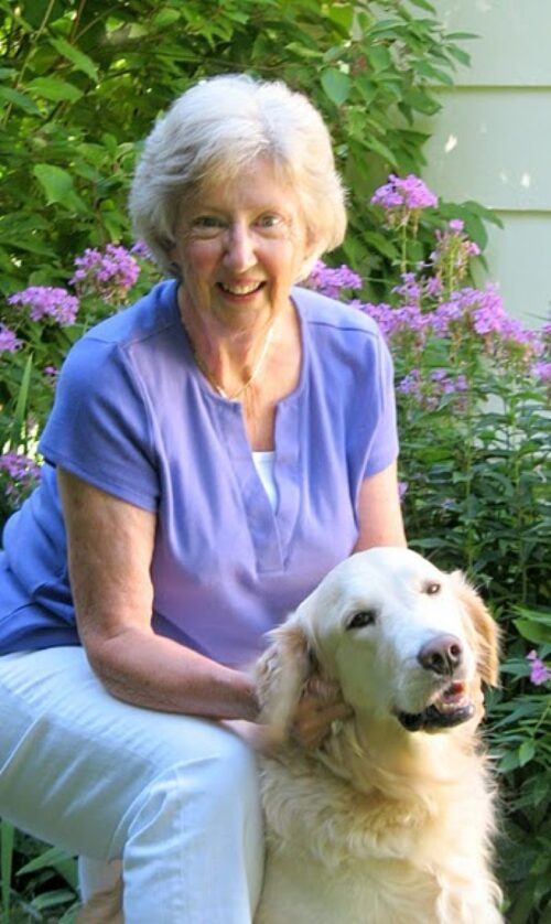 Janet Fritz sitting outdoors with her golden retriever in front of purple flowers