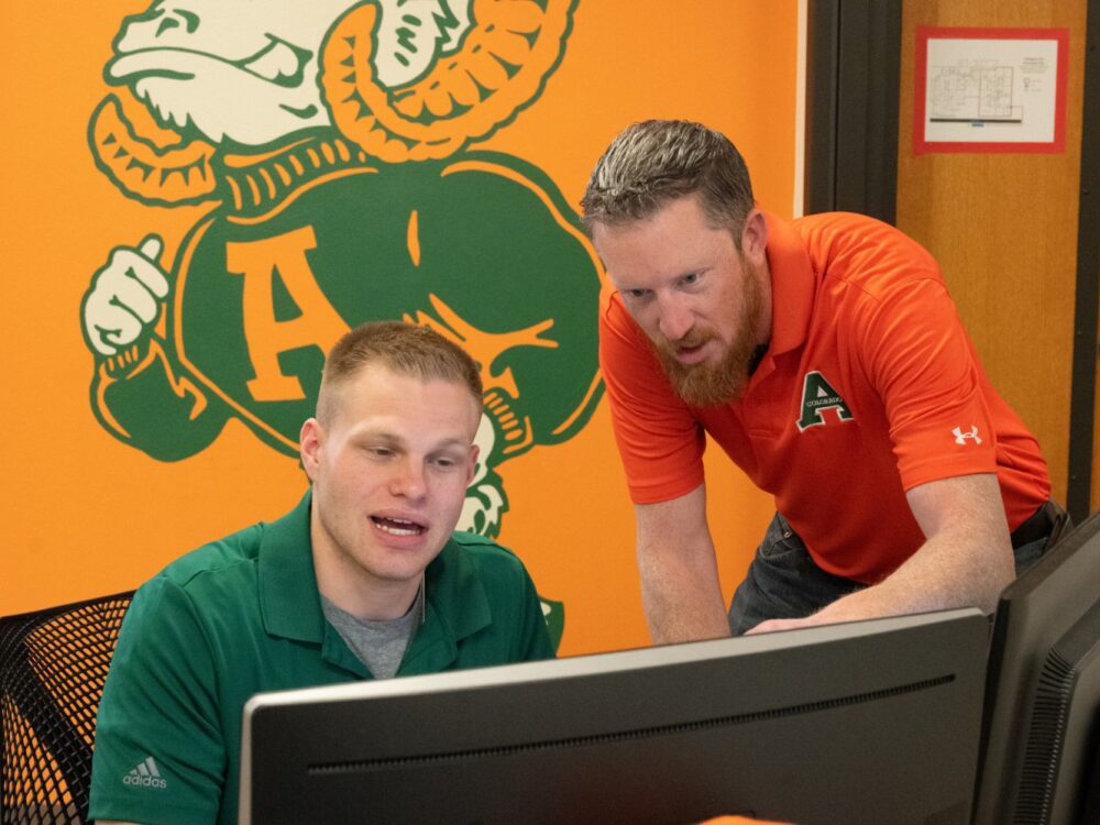 A professor wearing an orange Aggies shirt looking at a computer screen with a student