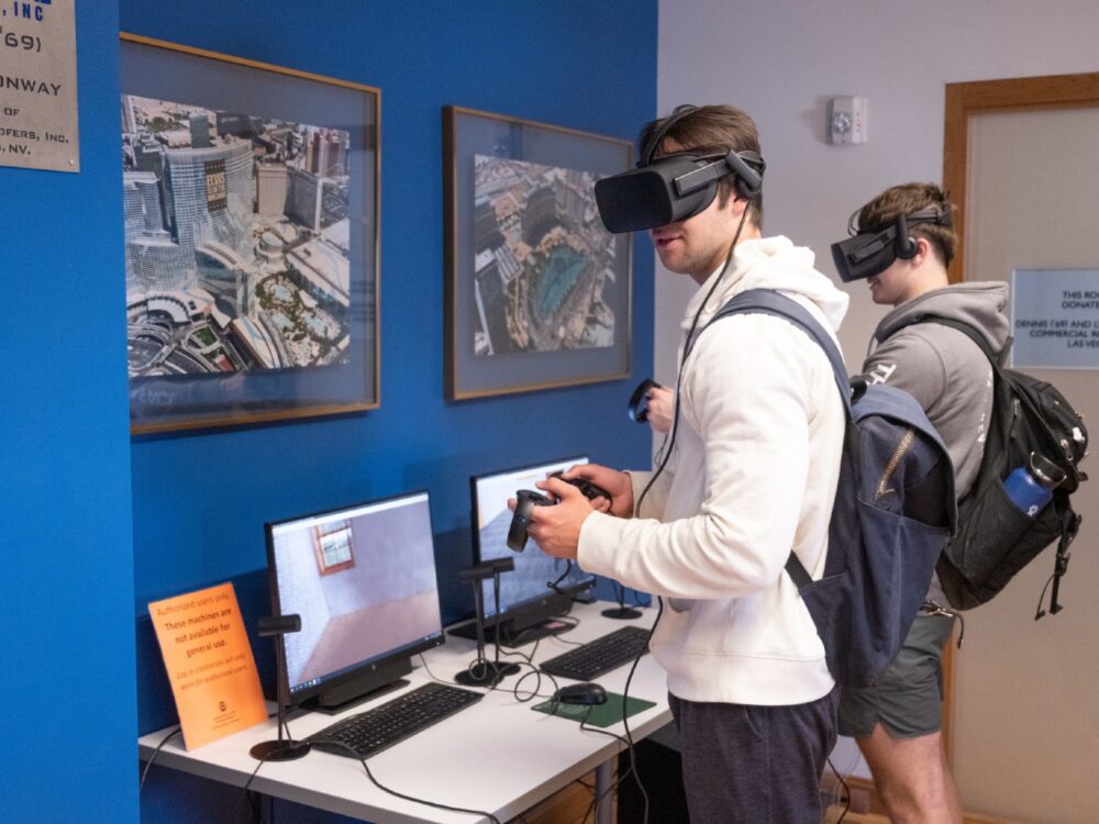 Two students wearing virtual reality goggles holding controllers and standing in front of computer screens