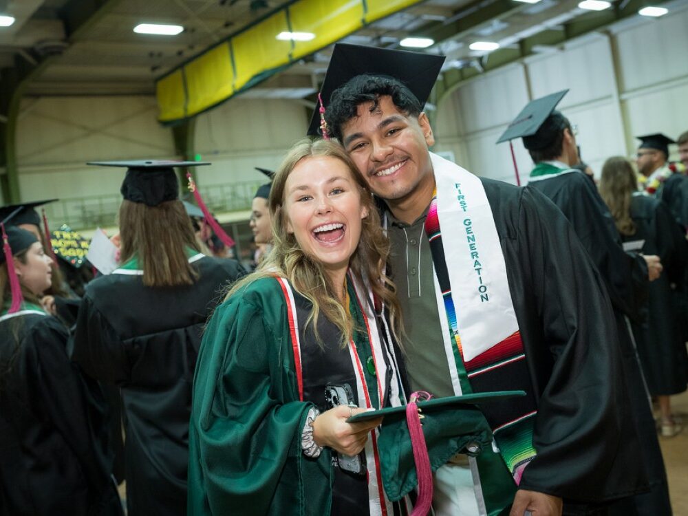 Two students standing together smiling in a room full of people wearing caps and gowns