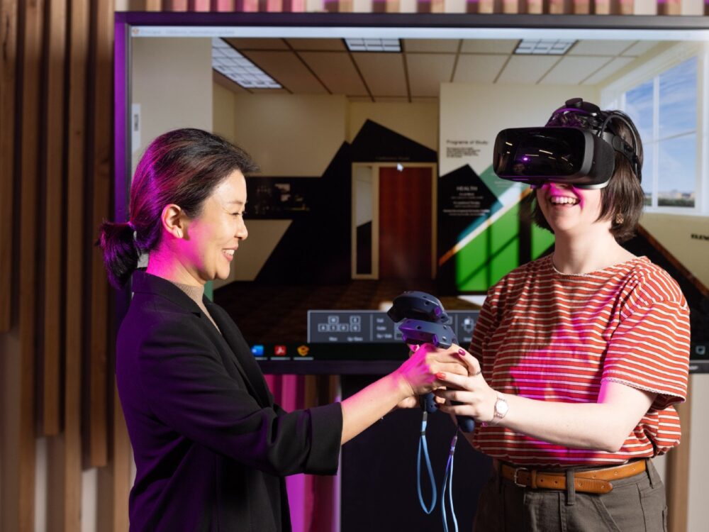 A faculty member handing controllers to a student wearing virtual reality goggles standing next to a large screen.