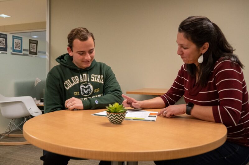 A student wearing a green CSU sweatshirt sits at a table with a career counselor looking at papers
