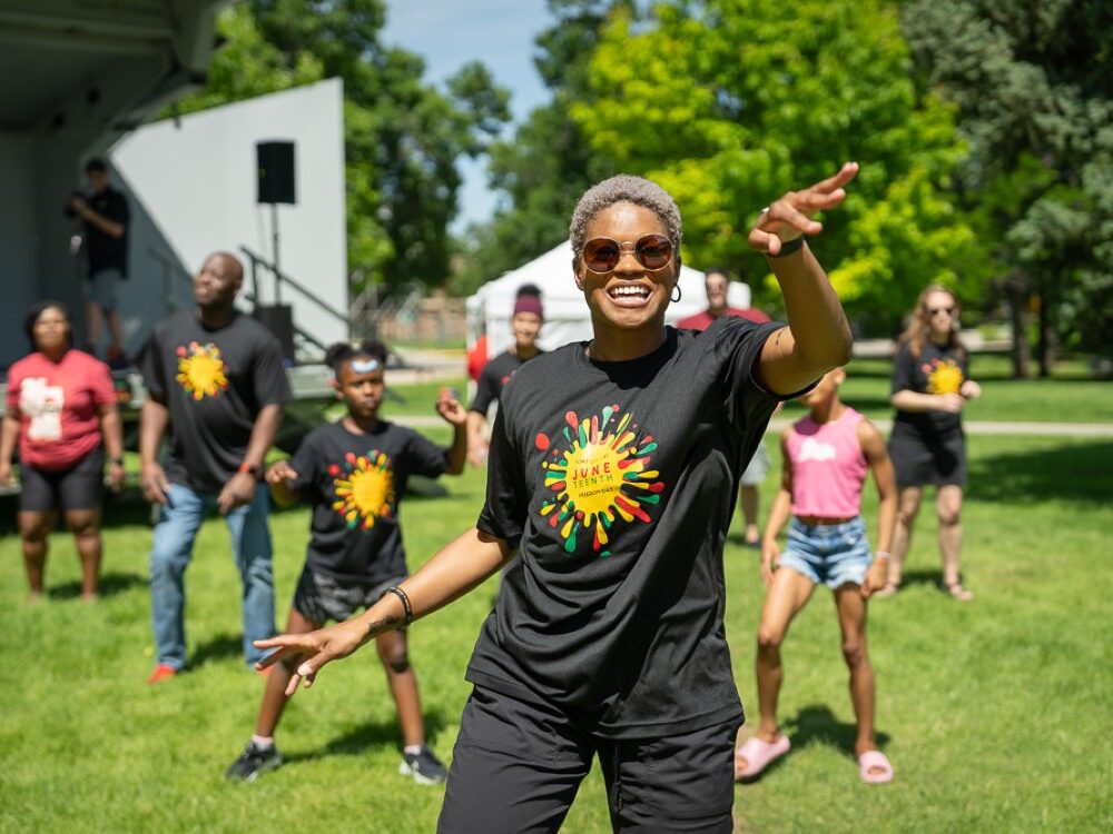A woman dances in a black Juneteenth t-shirt with children behind her.