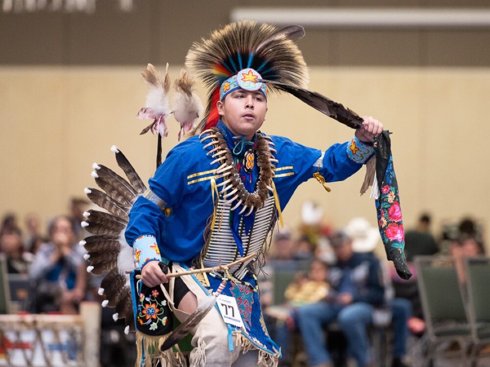 A Native American wearing traditional dress dances with drummers in the background