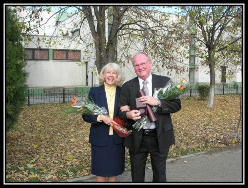 Vicky Buchan holding flowers standing next to her husband who is holding flowers and awards - outdoors and both are wearing suits