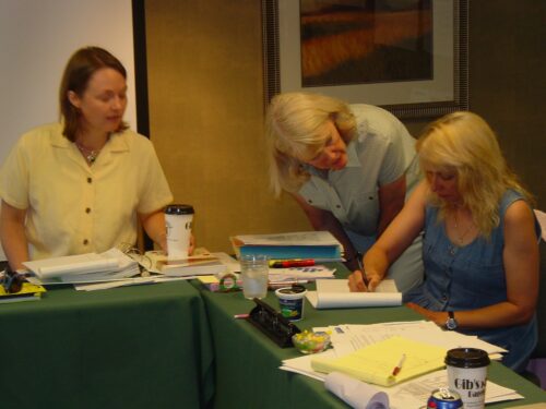 Vicky Buchan standing over a woman who is writing something on a paper while another woman looks on