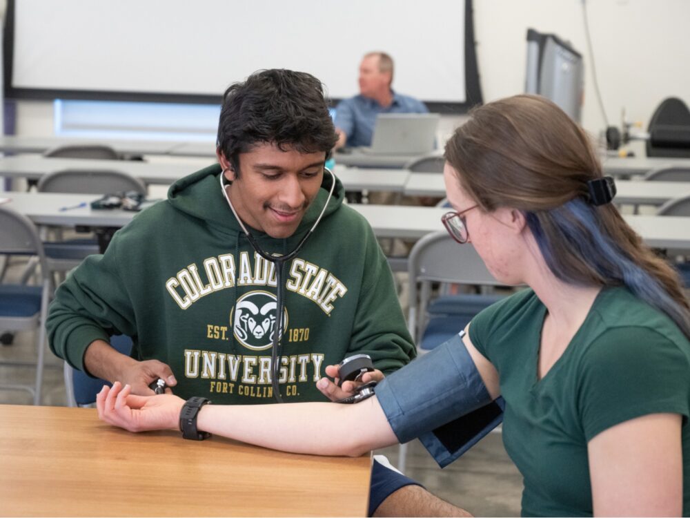 A student practices taking blood pressure with another student using a stethoscope and cuff in a classroom setting at Colorado State University.
