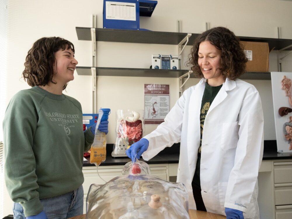 A professor wearing a white lab coat and gloves guides a student through a hands-on activity using a clear anatomical manikin in a classroom.