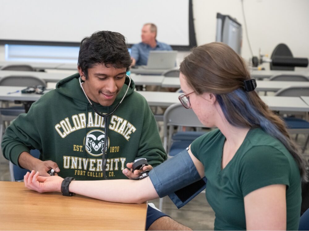 A student practices taking blood pressure with another student using a stethoscope and cuff in a classroom setting at Colorado State University.