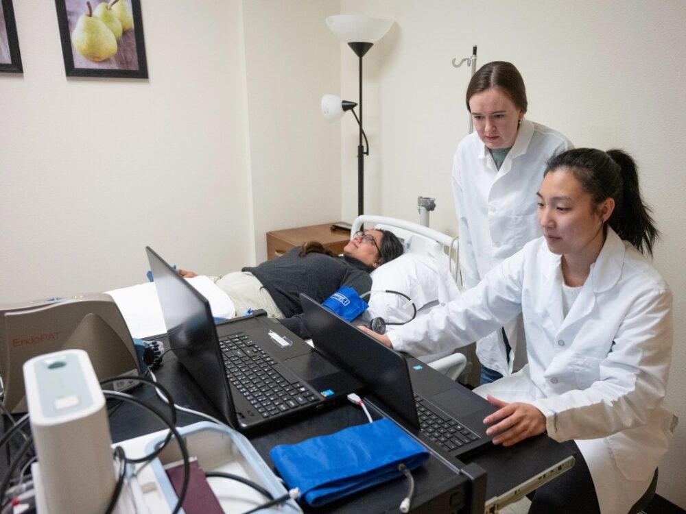 Two students in white coats look at a computer screen next to a patient in a hospital bed.