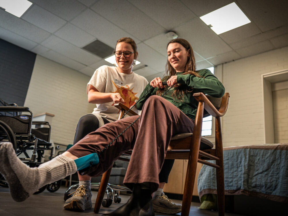 An OT student assists another student using an assistive device to pull on socks.