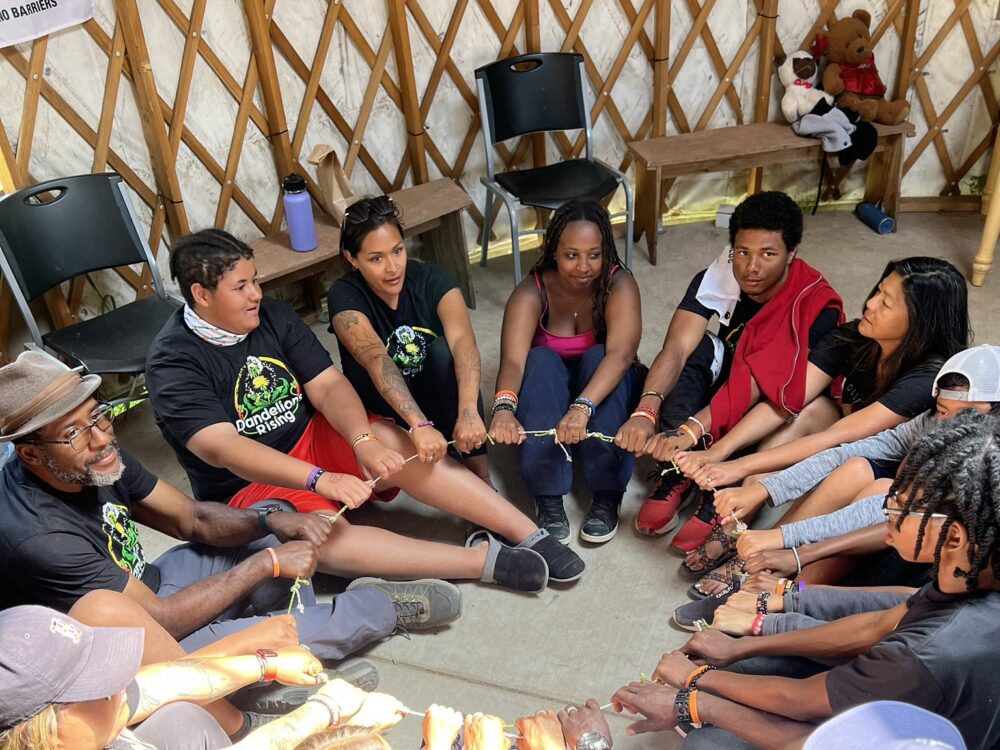 A group of BIPOC teens and adults sit in a circle, holding a hoop in the center.