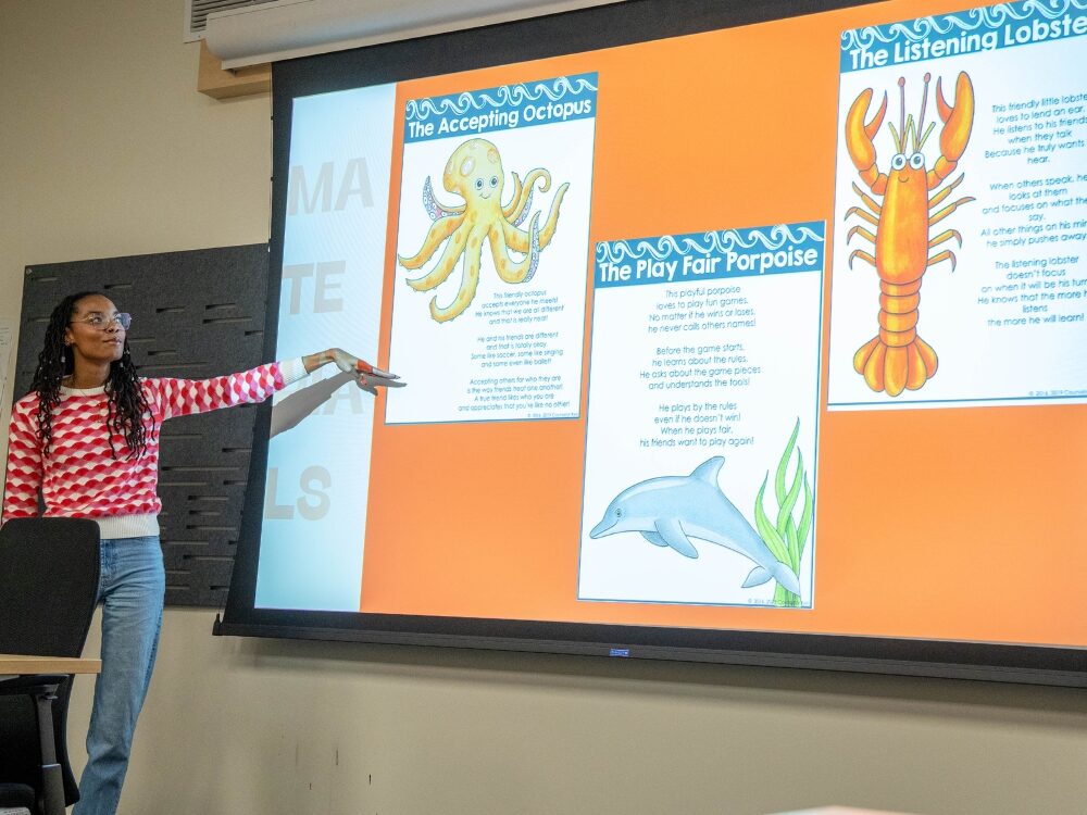 A CSU student smiles while teaching a class. She points to a screen that has various animal characters as part of a lesson.