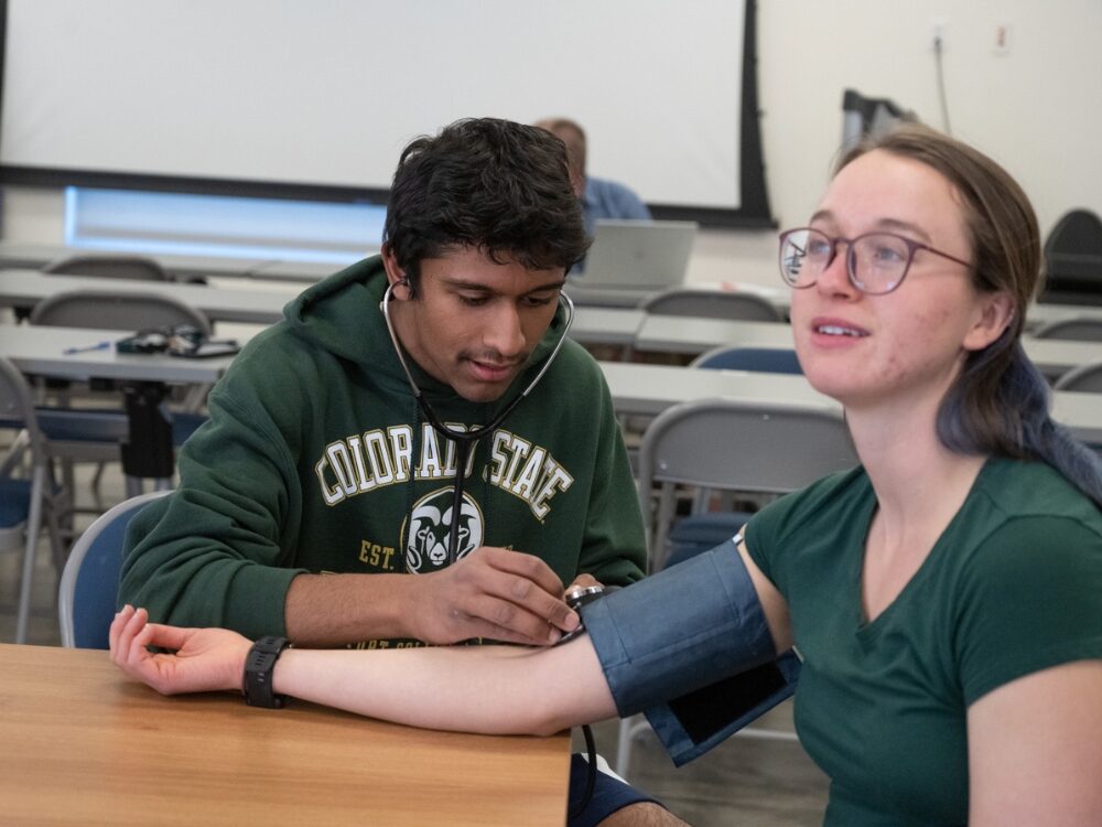 A student practices taking blood pressure with another student using a stethoscope and cuff in a classroom setting at Colorado State University.